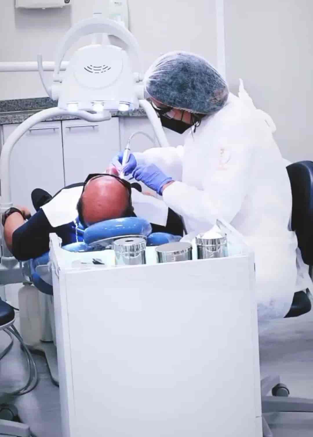 Denstista mulher sorrindo de jaleco branco atendendo um homem deitado em uma cadeira de dentista.