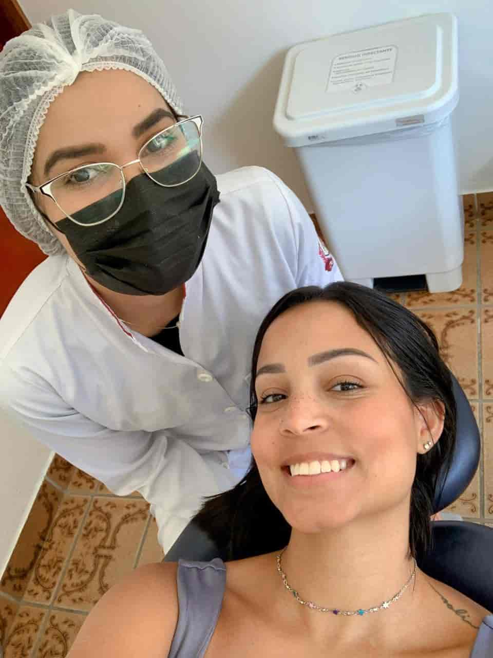 Duas mulheres uma Dentista de jaleco branco e mascara preta e oculos, a outra uma paciente de cabelo preto e sorrindo deitada em uma cadeira de dentista.